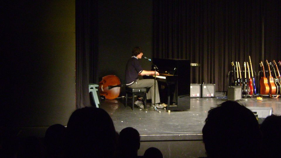 Photo is of Patrick performing City of Lights at his high school. He's wearing his school uniform, sitting at a black upright piano, on stage in front of the audience.